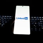 Hand holding smartphone with LinkedIn logo displayed on screen in front of illuminated keyboard in dark background.