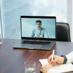 Excited developer in video interview on laptop during virtual tech job discussion, surrounded by notes and water glass on wooden desk.