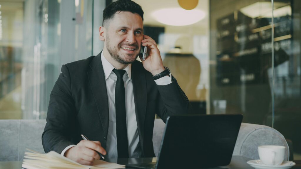 A professional real estate agent in a suit talking on his phone and taking notes in an office, demonstrating effective communication and active listening during a business negotiation.