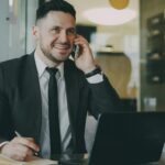 A professional real estate agent in a suit talking on his phone and taking notes in an office, demonstrating effective communication and active listening during a business negotiation.